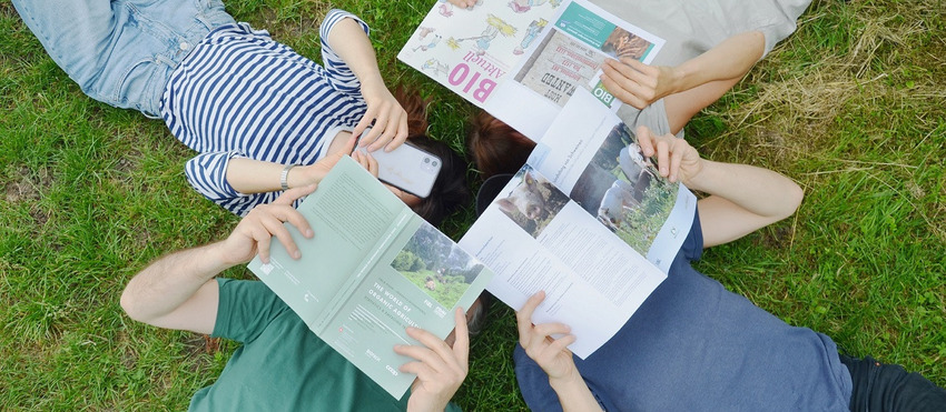 Vier Personen liegen auf dem Rücken im Gras und informieren sich über Zeitschriften beziehungsweise am Handy über landwirtschaftliche Themen.