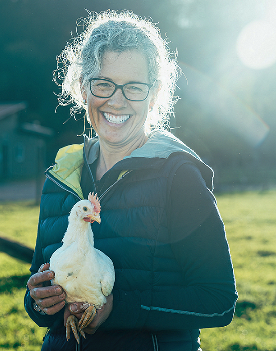 Ca. 50-jährige Bäuerin, die ein weisses Huhn hochhebt, strahlt in die Kamera. Wiese und audgehende Sonne im Hintergrund.