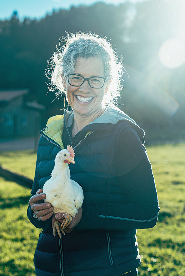 Ca. 50-jährige Bäuerin, die ein weisses Huhn hochhebt, strahlt in die Kamera. Wiese und audgehende Sonne im Hintergrund.